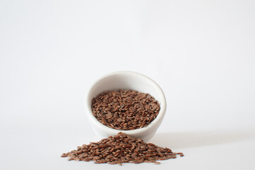 Flaxseeds, profile view of seeds spill from a small, white bowl onto a white surface in a minimalist scene with isolated background.