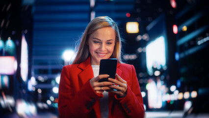 Beautiful Woman Standing, Using Smartphone on City Street with Neon Bokeh Lights Shining at Night. Confident Smiling Beautiful Female Using Mobile Phone. Medium Tracking Cinematic Portrait.