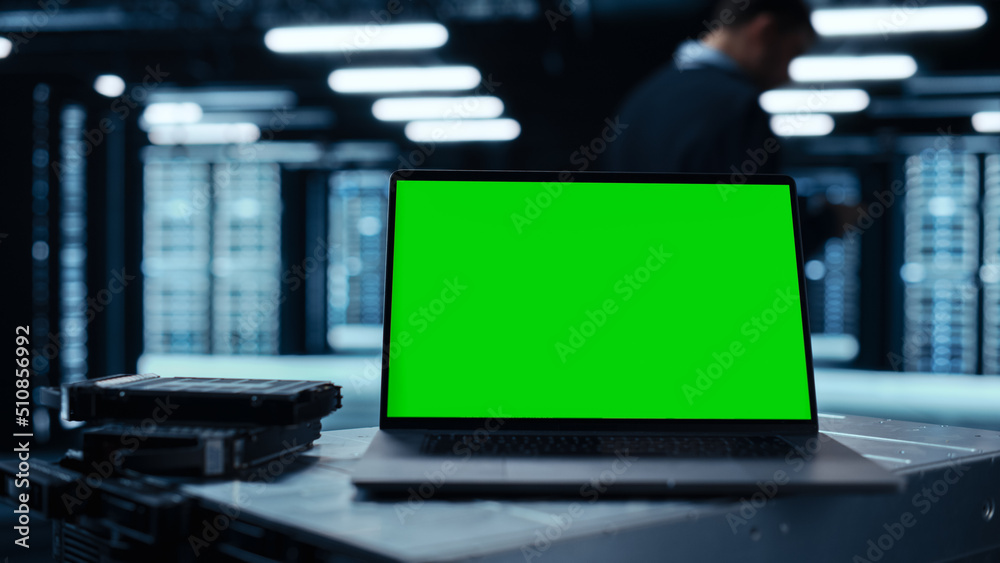 Poster Laptop Computer with Green Screen Chroma Key Mock Up Display Stands on a Table. In the Background Data Center With Rows of Server Racks and IT Engineer Working. Advanced Equipment.