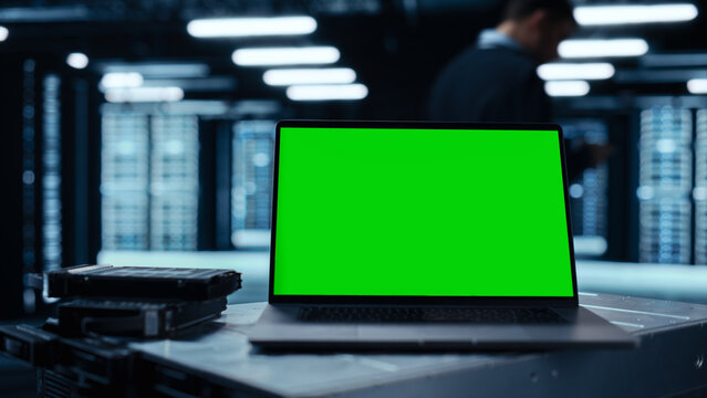 Laptop Computer with Green Screen Chroma Key Mock Up Display Stands on a Table. In the Background Data Center With Rows of Server Racks and IT Engineer Working. Advanced Equipment.