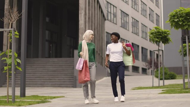 Two pretty multiracial young best friends women, walking in beautiful modern city, holding colorful shopping bags, having fun and laughing together.