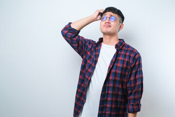 Handsome young Asian man wearing casual shirt and glasses standing with a shocked expression and holding his head