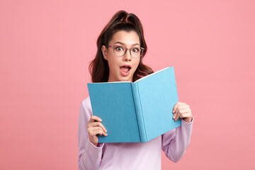 Cute Asian girl is holding the book and standing on pink background with surpirsed face, blank copy space for your advertising content.