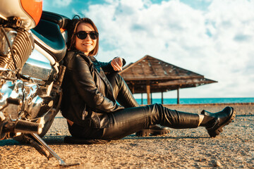 Independent happy woman in a leather motorcycle outfit poses sitting on the ground near a...