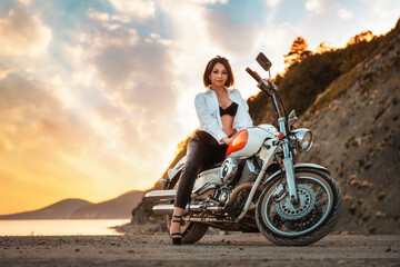 Obraz na płótnie Canvas Beautiful sexy adult woman with a high heels and leather pants, posing sitting on motorcycle. Epic sunset sky on the background. The concept of Motorcyclist Day and moto travel