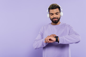 bearded african american man in wireless headphones looking at smart watch isolated on purple.