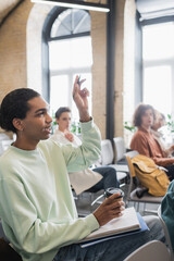 african american student with coffee to go and raised hand asking question during lecture in university.
