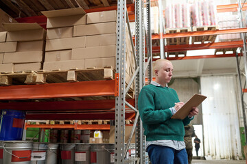 Female warehouse worker making notes in document to control number of goods on shelves during...