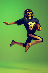 Fototapeta na wymiar Sportive little boy in sports uniform and equipment playing american football isolated on green background in neon light. Concept of sport, movement, achievements.