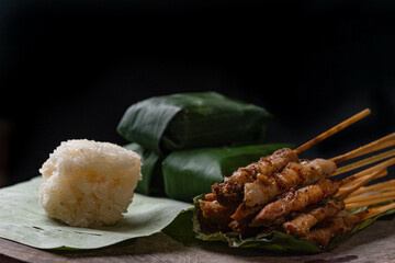 front view of grilled skewered pork(Moo Ping) with white sticky rice and sticky rice wrapped in banana leaves placed on the banana leaves and wood floor, black background