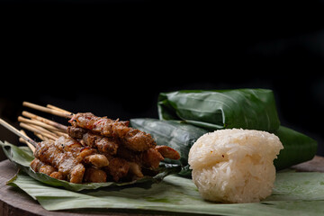 front view of grilled skewered pork(Moo Ping) with white sticky rice and sticky rice wrapped in banana leaves placed on the banana leaves and wood floor, black background