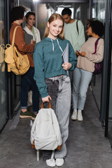 happy woman with copybook and backpack looking at camera near blurred interracial students.