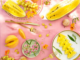 Sunny and bright open air breakfast with yellow watermelon, grape, plums and summer wild flowers decoration. Romantic picnic for couple. Floral botanical background, pattern.