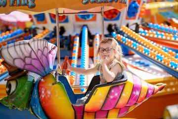 Adorable little preschool girl with glasses riding on animal on roundabout carousel in amusement...