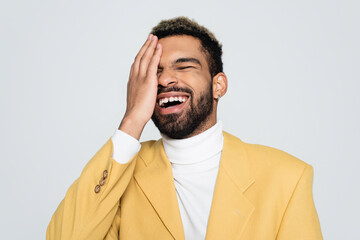 happy african american man in pastel yellow blazer closing eye and laughing isolated on grey.