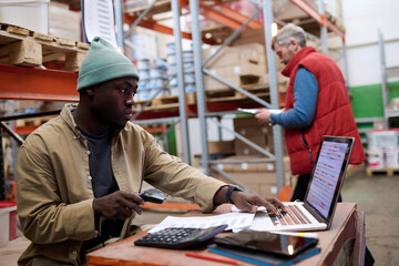 African American distributor using laptop to scan barcodes of parcels while sitting at his...