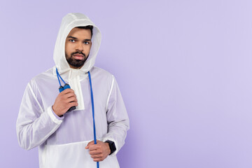 african american sportsman in stylish sportswear with hood looking at camera and holding skipping rope isolated on purple.