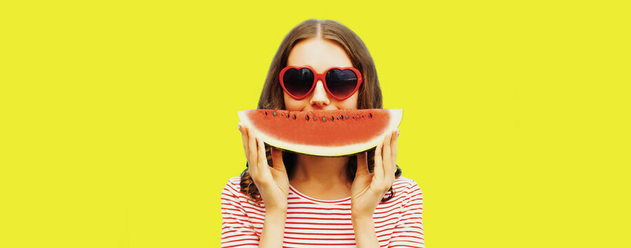 Portrait of happy smiling young woman with slice of watermelon wearing red heart shaped sunglasses on yellow background, blank copy space for advertising text