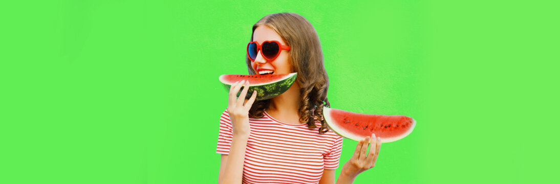 Portrait of happy smiling young woman with slice of watermelon wearing red heart shaped sunglasses on green background, blank copy space for advertising text