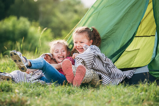 Two little girls sisters sitting on the green grass next to camping tent entrance, cheerfully laughing about jokes they told to each other. Careless childhood and outdoor activities concept photo.