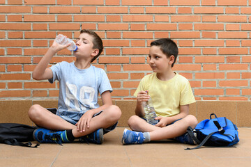 Boys sitting on the ground drinking water from a plastic bottle next to his school bag.