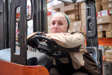 Portrait of female driver of forklift sitting behind the wheel and looking at the camera during her work at warehouse
