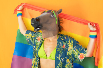 Happy gay woman in horse head mask holding LGBT rainbow flag isolated on orange background