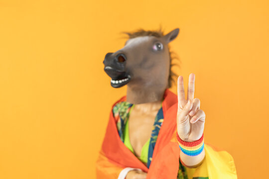 Happy gay female in surreal horse head mask showing the victoria sign gesture ,wrapped with LGBT rainbow flag isolated on orange background