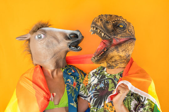 Portrait of funny gay couple wearing horse and dinosaur animal mask wrapped with LGBT pride rainbow flag isolated on yellow studio yellow background.Surreal people friends pride gay concept.