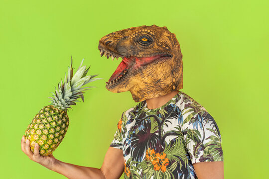 Portrait of happy man wearing dinosaur and hawaiian shirt holding pineapple fruit in hand.Funny celebration with copy space isolated on studio green background.Tropical travel destination 