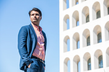 Businessman in shirt and jacket outdoors. Hands in the pocket. Behind him a building and the blue...