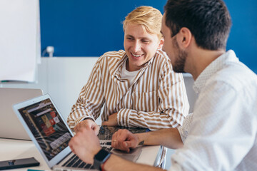 Two men looking at laptop screen while sitting at desk in office