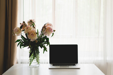 Spring home office still life composition. Laptop and vase with bouquet white pink peonies flowers on a table. Cozy home office
