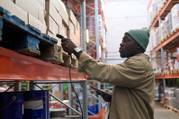African manager scanning packages on shelves with special device during his work at warehouse