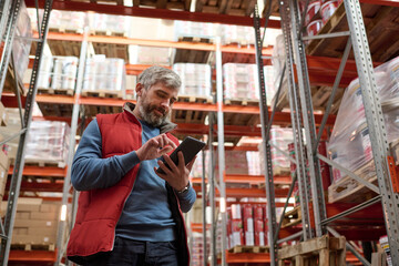 Fototapeta na wymiar Mature warehouse worker using digital tablet for checking stock on shelves standing opposite cardboard boxes