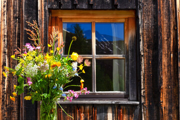 Fototapeta na wymiar altes Fenster eine Almhütte mit Spiegelungen der Berge im Fenster, davor steht ein bunter Blumenstrauß 
