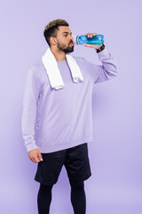 bearded african american man in sweater standing with white towel and drinking water isolated on purple.