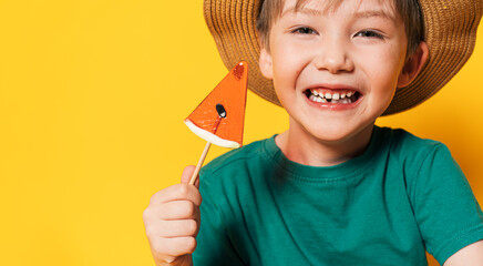 Boy in hat holding lollipop in the form of piece of watermelon in his hand against yellow...