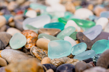 Natural polish textured sea glass and stones on the seashore. Azure clear sea water with waves. Green, blue shiny glass with multi-colored sea pebbles close-up. Beach summer background