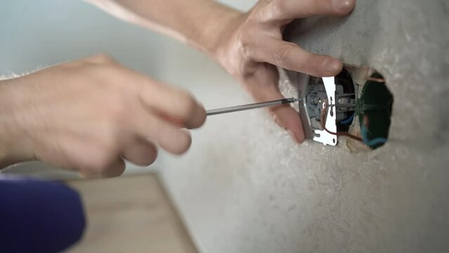 Household husband, electrician, repairs, installs a socket in the room. The hand of the master twists the socket with a screwdriver close-up on background of the wall.