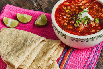traditional mexican food, beef pancita, menudo with green chili, onion and lemon with freshly made tortillas on wooden table and a pink mexican napkin