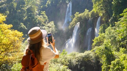 Happy female tourist with backpack in travel. Wanderlust, tourism concept. Young woman traveler make vertical video for social media of amazing waterfalls and tropical nature on her smartphone camera.