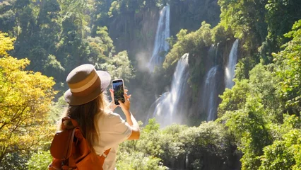 Foto op Aluminium Happy female tourist with backpack in travel. Wanderlust, tourism concept. Young woman traveler make vertical video for social media of amazing waterfalls and tropical nature on her smartphone camera. © TravelMedia