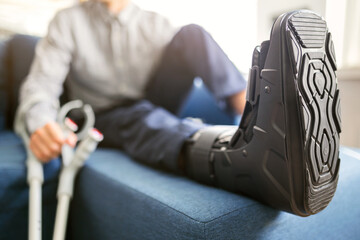 Man with ankle orthosis, black splint, walker boot on leg sitting on the sofa at home. Ankle foot orthosis close-up.. - obrazy, fototapety, plakaty