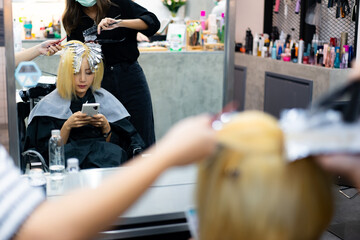 Mirror reflection from hairdressing table of young short-haired Asian female client sitting on hairdressing chair using a cell phone while having a hair dyeing by Asian female hairdressers in salon.