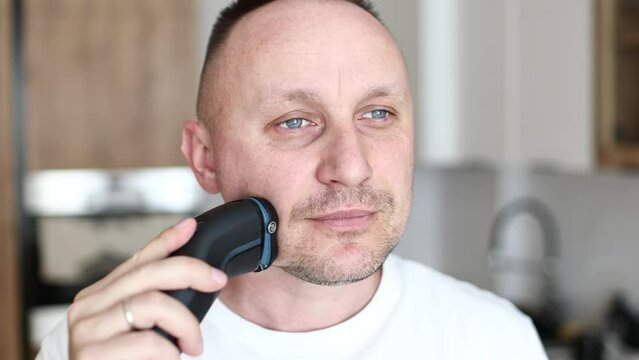 Handsome young unshaven man shaving with modern electric razor trimer while looking at camera, indoors. Daily routine male self-care. man shaves with an electric razor in the kitchen.