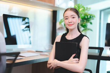 Portrait of successful and smiling young business woman holding file feeling confident and proud while standing in office and looking at camera