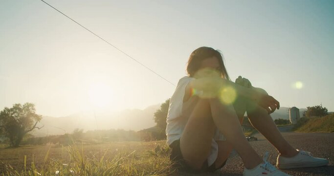 Attractive woman sit on meadow side at sunset after ride on skateboard. Hipster girl or teenager rest after active leisure and healthy lifestyle. Female enjoying fresh summertime air