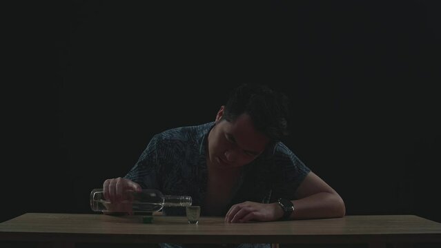 Drunk, Depressed Asian Man Pouring Vodka In A Shot Glass Before Drinking And Using A Fist Smashing The Table In Black Background
