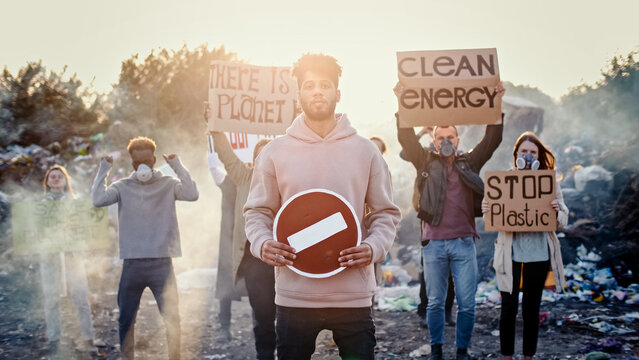 Portrait of Attractive Young Man Activist Holding Stop Sign. In Background Fighting People Protesting Against Garbage Pollution Staying at Dump in City Outskirts.